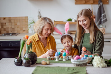 Happy easter family elderly grandmother and young woman mother blonde with little daughter with rabbit ears are preparing for the holiday to paint eggs