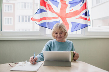 Senior woman writes new English words during distant learning with laptop, language classes for Ukrainian refugee for better adaptation abroad, British flag on the window, social distance education