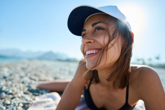 Cheerful young woman in white cap lying down on sunny beach and looking away during summer holiday.