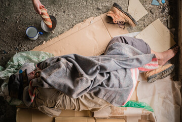 Old man beggar homeless wears sweater with blanket sleeping on cardboard at shelter with bowl for...