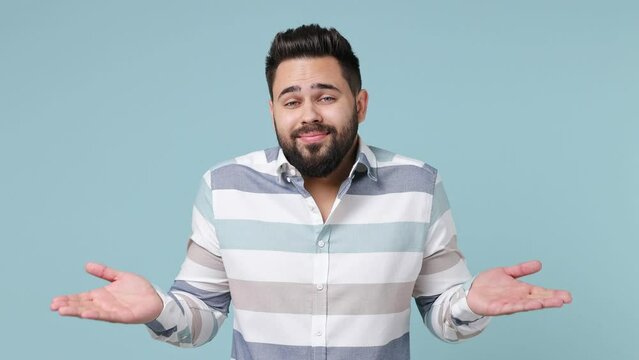 Fun confused shy shamed young bearded brunet man 20s wears striped shirt look camera spreading hands say oops ouch oh omg i am so sorry isolated on plain pastel light blue background studio portrait