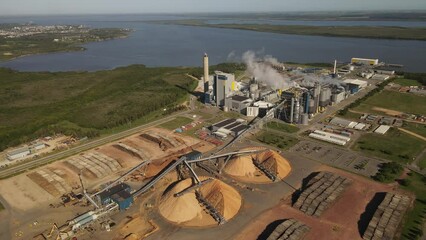 Aerial view of Paper Mill Factory with smoky chimneys front to River - Sawdust piles and pipes transporting wood trunks