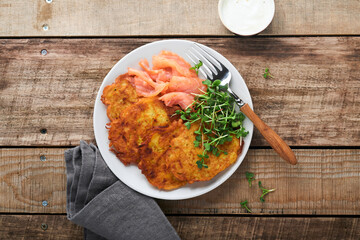 Potato pancakes. Fried homemade potato pancakes or latkes with cream, green onions, microgreens, red salmon and sauce in rustic plate on old wooden table background. Rustic style. Top view.
