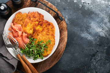 Potato pancakes. Fried homemade potato pancakes or latkes with cream, green onions, microgreens, red salmon and sauce in rustic plate on old black table background. Rustic style. Top view.
