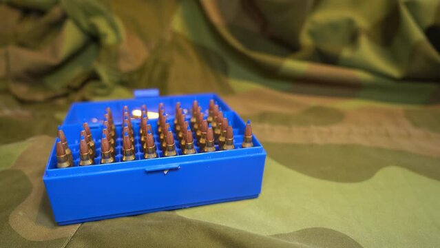 Ammunition box full of powerful military grade 223 caliber varmint bullets with blue plastic tip laying on camouflage jacket - Slider from left to right