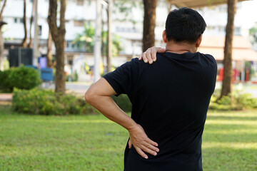 Fototapeta premium Back view of man hurts his shoulder and waist at outdoor park. Concept : Health problems, swelling and inflammation, pain. Symptoms of health injury in daily life. 