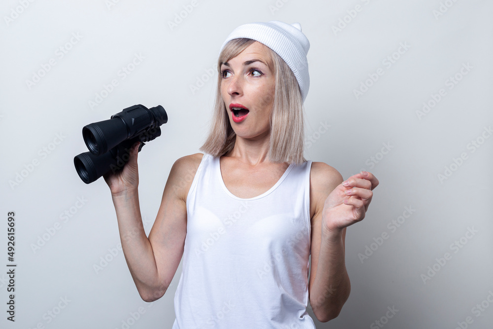 Wall mural Surprised young woman in a white hat with binoculars on a light background