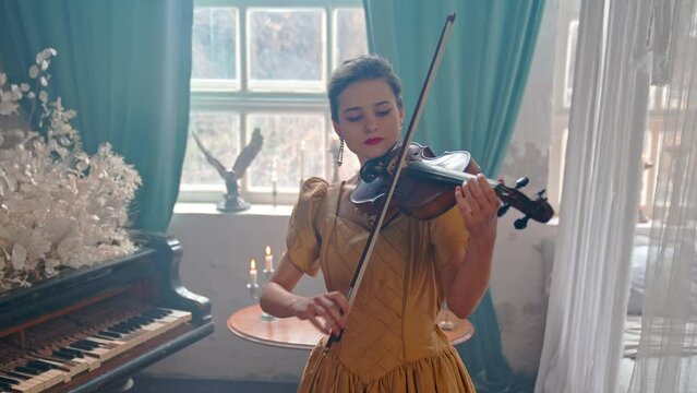 A Young Girl And Man Plays The Violin In A Beautiful Retro Room With Flowers And A Piano.