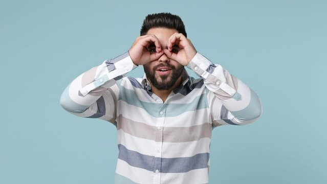 Smiling happy funny young bearded brunet man 20s years old wears striped shirt holding hands near eyes imitating glasses or binoculars isolated on plain pastel light blue background studio portrait