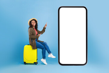 Happy young black woman sitting on suitcase and pointing at smartphone with mockup for online...