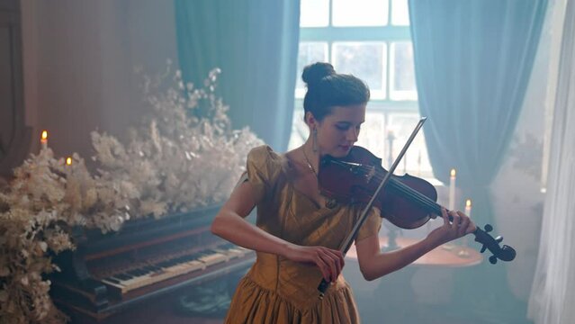 A Young Girl And Man Plays The Violin In A Beautiful Retro Room With Flowers And A Piano.