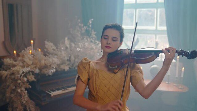 A Young Girl And Man Plays The Violin In A Beautiful Retro Room With Flowers And A Piano.
