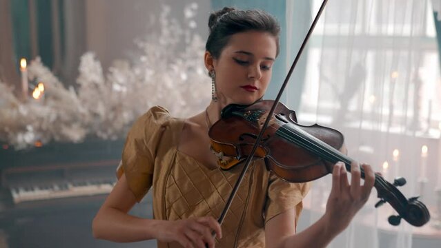 A Young Girl And Man Plays The Violin In A Beautiful Retro Room With Flowers And A Piano.