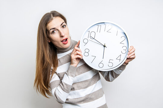 Surprised young woman holding white wall clock on light background.