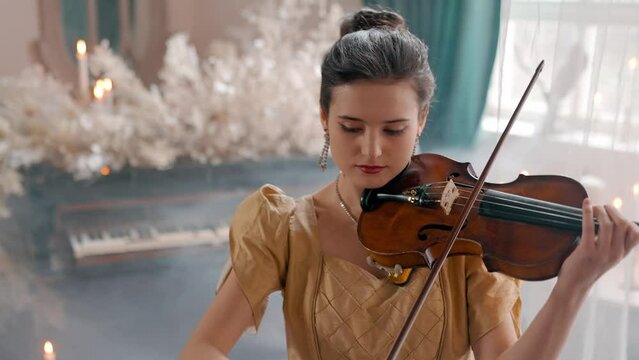 A Young Girl And Man Plays The Violin In A Beautiful Retro Room With Flowers And A Piano.