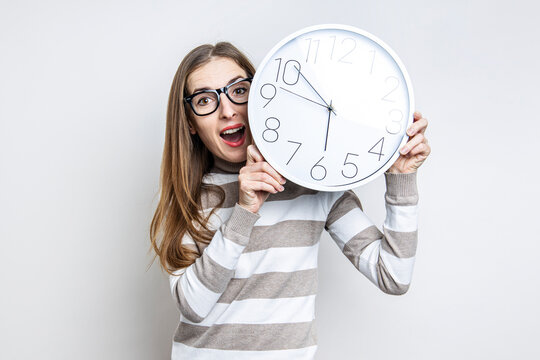 Surprised young woman holding white wall clock on light background.