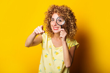 smiling young woman pointing her finger looks through a magnifying glass on a yellow background.