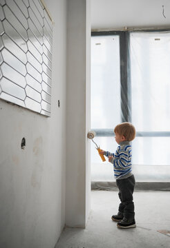Side view of cute little boy standing with special brush for repair and painting wall in white color. Concept of drawing wall at home in empty room for modern repair.