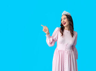 Beautiful young woman in stylish dress and tiara pointing at something on blue background