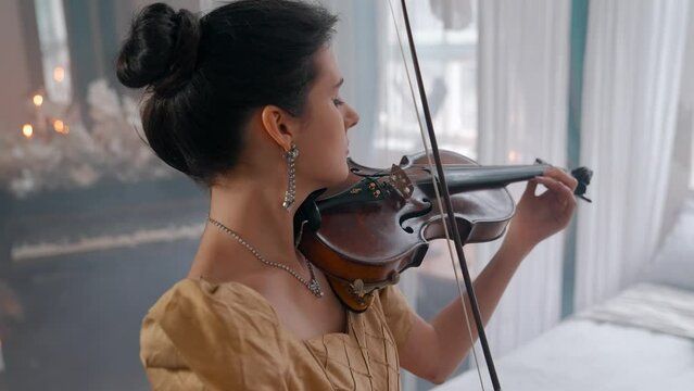 A Young Girl And Man Plays The Violin In A Beautiful Retro Room With Flowers And A Piano.