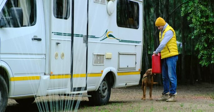 A man in a yellow vest and an autumn hat refuels a Caravan or motor home trailer in the yard of a private house before traveling. Preparing the car for a long journey. man with a dog in the yard