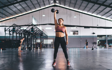 Determined Caucasian female in sportive wear standing in gym working on hands muscles using dumbbell, strong professional woman lifting weight - training upper arms biceps for keeping body in tonus