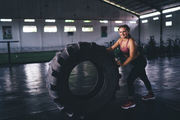 Fototapeta na wymiar Full length portrait of cheerful Caucaisan bodybuilder in tracksuit smiling at camera during time for weightlifting heavy wheel, successful female with sportive equipment posing during workout