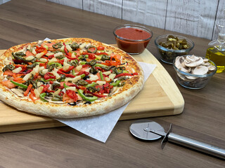 Not cut veggie pizza on wood board on table and pizza cutter and fresh mushrooms and olive oil on...