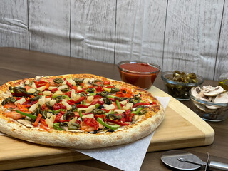 Not cut veggie pizza on wood board on table and pizza cutter and fresh mushrooms and olive oil on...