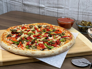 Not cut veggie pizza on wood board on table and pizza cutter and fresh mushrooms and olive oil on...
