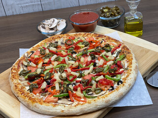 Not cut veggie pizza on wood board on table and pizza cutter and fresh mushrooms and olive oil on side.