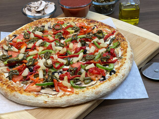 Not cut veggie pizza on wood board on table and pizza cutter and fresh mushrooms and olive oil on side.