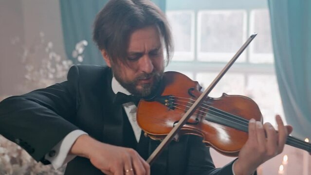 A Young Girl And Man Plays The Violin In A Beautiful Retro Room With Flowers And A Piano.