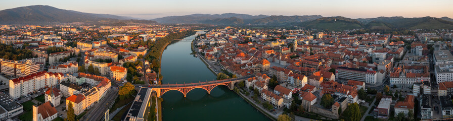 Fototapeta na wymiar Panoramic aerial view of Maribor, Slovenia early in the morning. Red bridge crossing river in a peaceful town in Europe. Many houses with red roofs from drone view.