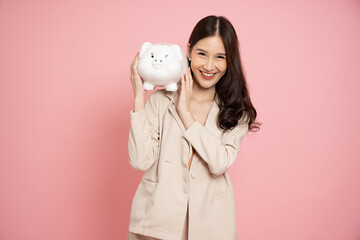 Portrait of Happy Asian businesswoman holding white piggy bank isolated on pink background, Saving...