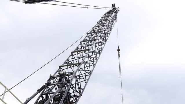 Looking up at the intricate structure of the boom of a crane with outer weights, jib hook, uffing wire rope, pulleys, and boom pendants. Sun trying to peak through the clouds in the sky. 