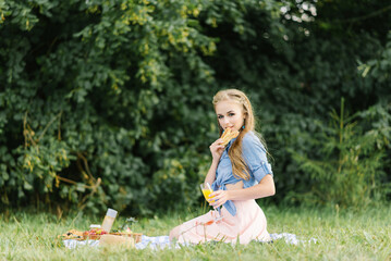 A smiling beautiful girl holds a fresh bun in her hands and sits on a blanket in the open air in the park. Summer picnic in the park