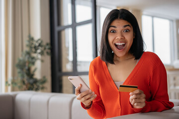 Excited African American woman holding credit card using mobile phone shopping online. Happy freelancer receive payment, check card balance. Emotional female ordering food online looking at camera