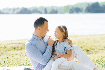 A happy father holds his daughter in his arms. They have fun in nature, sitting on a blanket by the lake in summer