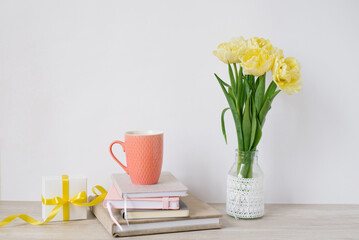 A mug, a lot of notepads in a stack, a bouquet of yellow spring tulips and a gift box on the desktop of a freelancer or blogger woman. Women's day, valentine's day or birthday. Front views