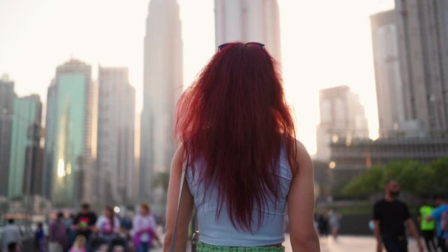 View from the back, a woman with red hair is walking in the center of Dubai against the backdrop of skyscrapers. United Arab Emirates. Slow motion. 