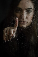 Blurred by the droplets - Beautiful long haired young woman pointing her index finger on a glass plate covered with droplets in front of her face, focus on the finger and droplets