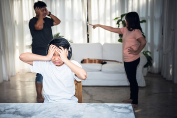 Obraz premium Asian teenage boy wearing headphones covering his ears with hands and stressed, with blur background of father and mother arguing, to people and family problems caused by quarrels concept.