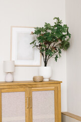An artificial flower in a pot on a wooden chest of drawers in the living room, decorative items and a frame on the wall in a Scandinavian minimalist style