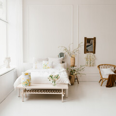 A bed in the bedroom with white linens, spring yellow flowers in a vase on the bedside table in the white interior of the bedroom
