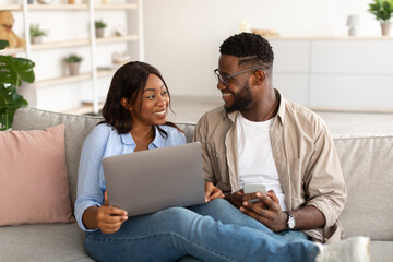 Fototapeta premium Happy black couple sitting on couch, using cellphone and pc