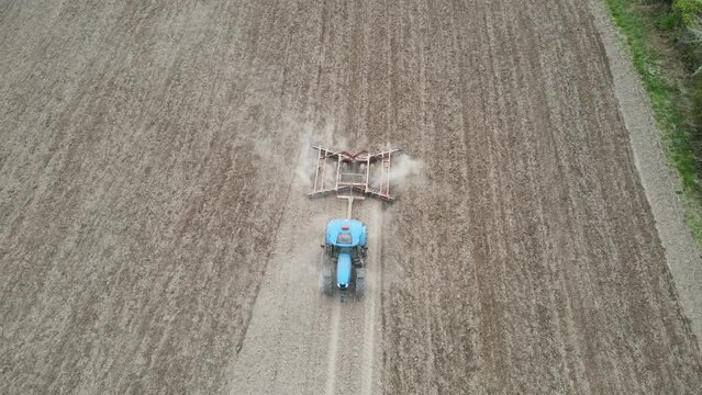 Birds eye view of farmer tilling field in preparation for planting. Evenly spaced rows in the dry soil kicking up dust. Tree line along the edge of the field. Blue tractor in the field. 