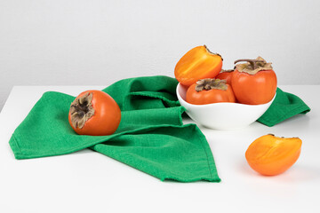 Still life with persimmon and green cloth on a white table. The fruits are in a white bowl. Horizontal orientation.