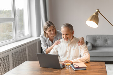 Old man and an age woman sit at home at a table in a laptop. Charming woman and gray-haired man with a beard surf on the Internet.