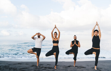 Young man and woman in sportive clothes standing in yoga pose breathing and enjoying recreation outdoors, group of diverse yogi feeling inspiration and balance in asana training at seashore beach